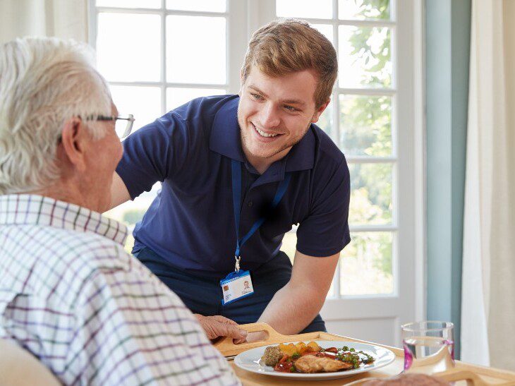 senior man smiles at his caregiver while enjoying a hearty meal at home