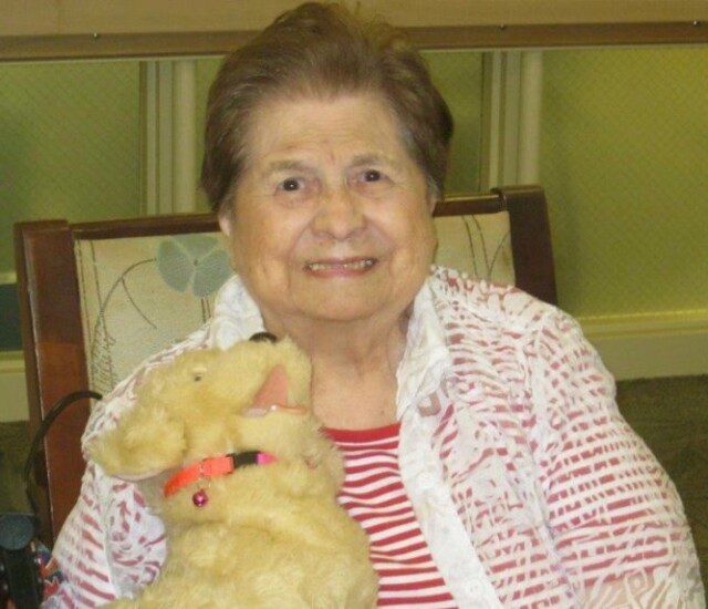 smiling senior resident seated in chair holds stuffed dog and looks at the camera