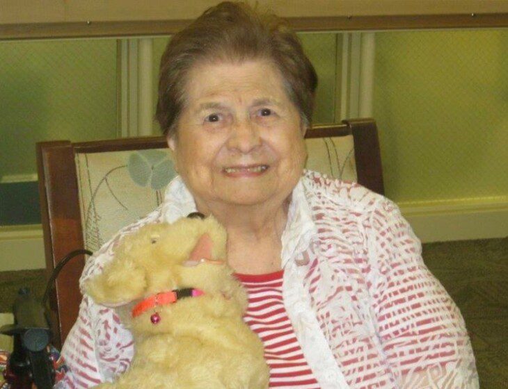 smiling senior resident seated in chair holds stuffed dog and looks at the camera