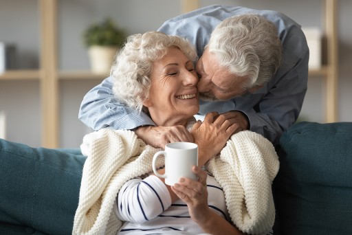 senior man kisses his wife's cheek as she sits on a couch