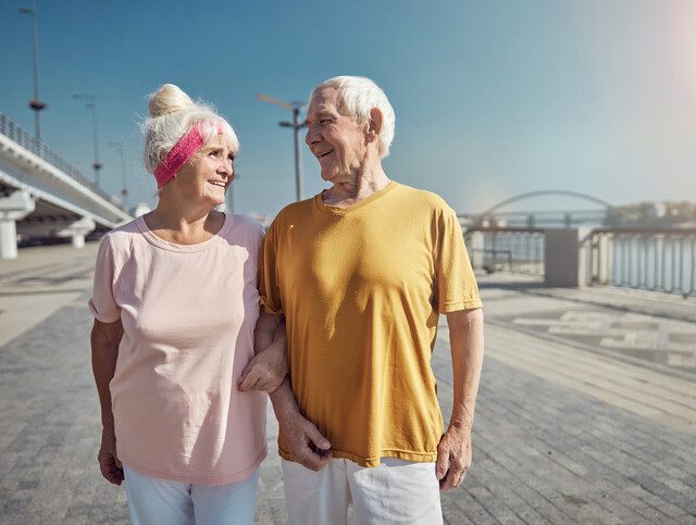 senior couple strolls arm-in-arm together, smiling, along a waterfront boardwalk