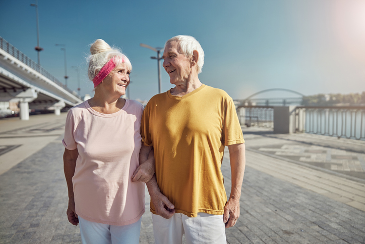 senior couple strolls arm-in-arm together, smiling, along a waterfront boardwalk