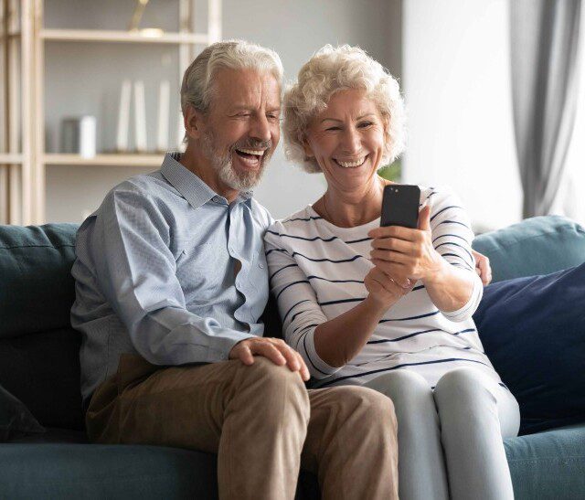 seated on a couch, a senior couple smiles and looks at their phone screen for a video call