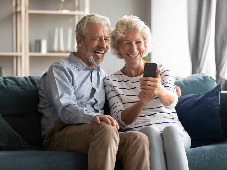 seated on a couch, a senior couple smiles and looks at their phone screen for a video call