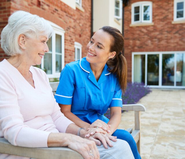 senior woman and her caregiver sit on a bench outside, smiling and conversing