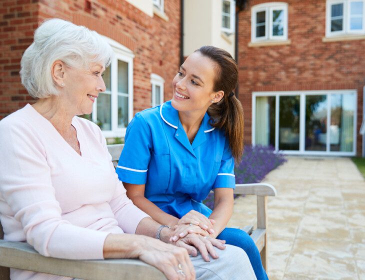 senior woman and her caregiver sit on a bench outside, smiling and conversing