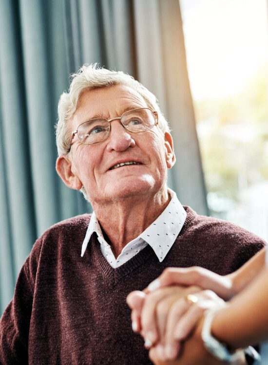 seated senior man smiles up at his caregiver, clasping her hand