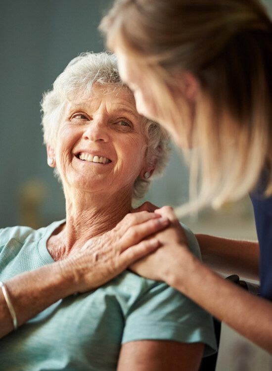 seated senior woman smiles at her female caregiver, who places her hand gently on her shoulder