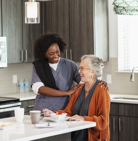 senior woman seated at the island in her home smiles while enjoying breakfast in the company of her caregiver