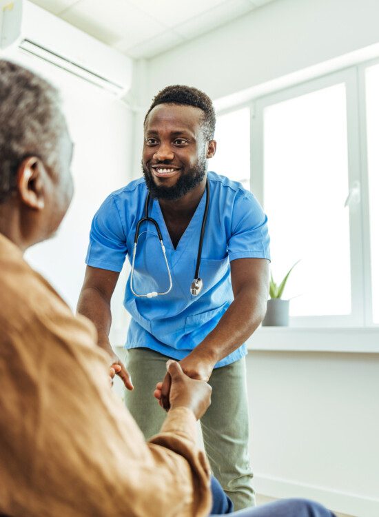 seated senior man gets up from a chair with the assistance of his smiling caregiver