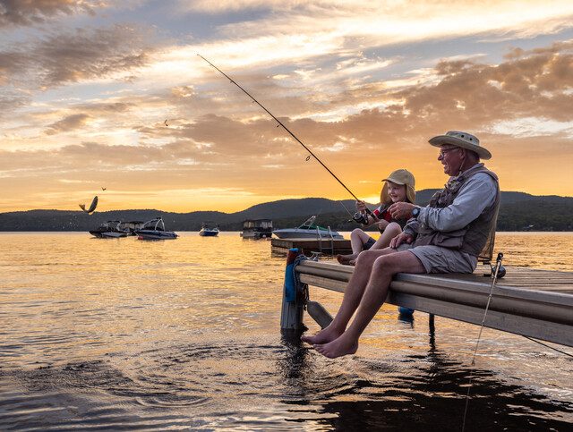 senior man and his grandson sit on a small fishing pier at a pond, smiling and fishing in the sunset