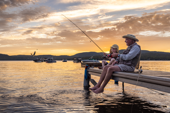 senior man and his grandson sit on a small fishing pier at a pond, smiling and fishing in the sunset
