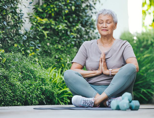 senior woman sits on yoga mat outdoors meditating, dumbbell weights beside her