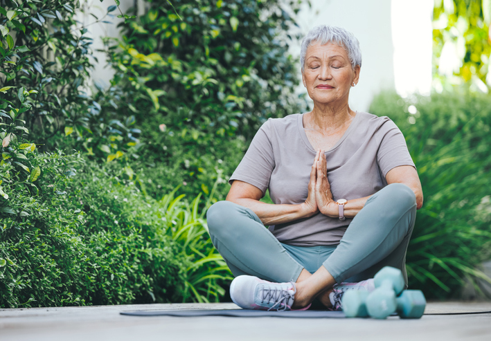 senior woman sits on yoga mat outdoors meditating, dumbbell weights beside her