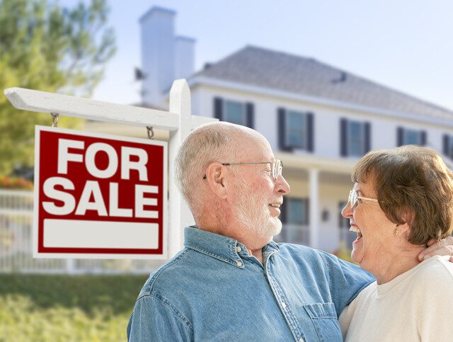 senior couple embraces and smiles in front of their home with a "for sale" sign, celebrating