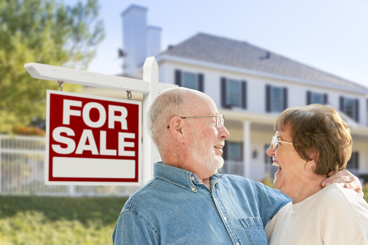 senior couple embraces and smiles in front of their home with a "for sale" sign, celebrating