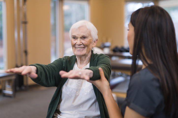 seated senior woman performs physical therapy exercise with the help of her trainer