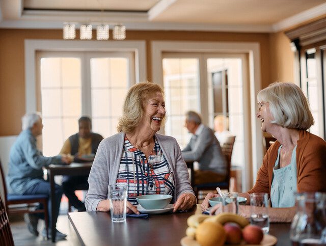 two senior women laugh while enjoying breakfast together indoors at Village on the Green Senior Living Community