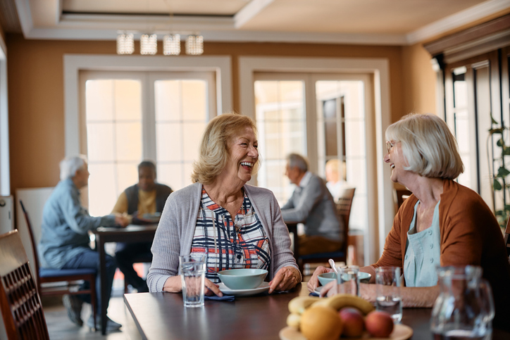 two senior women laugh while enjoying breakfast together indoors at Village on the Green Senior Living Community