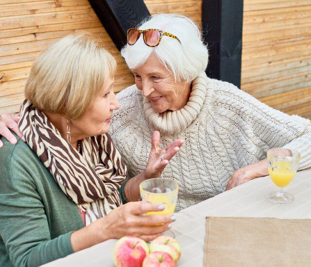 two senior women lean in to speak conspiratorially while smiling and enjoying mimosas