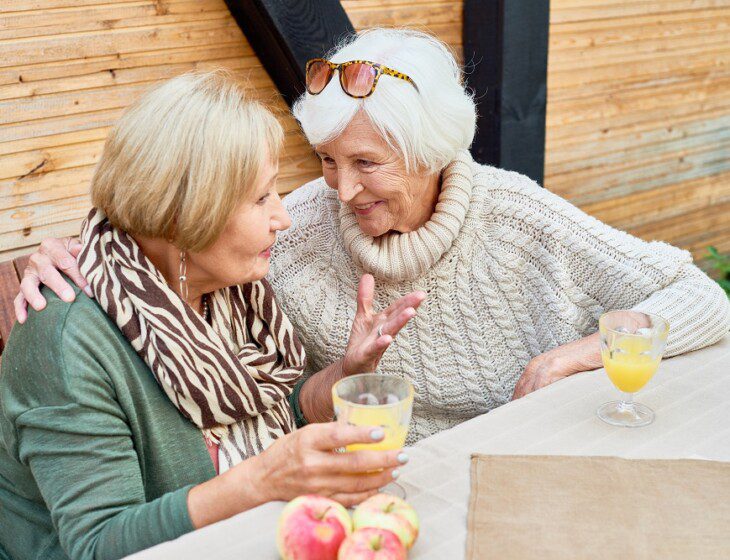 two senior women lean in to speak conspiratorially while smiling and enjoying mimosas