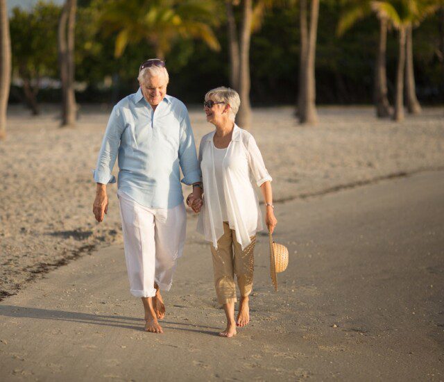 smiling senior couple holds hands and walks facing the camera as they leave the beach