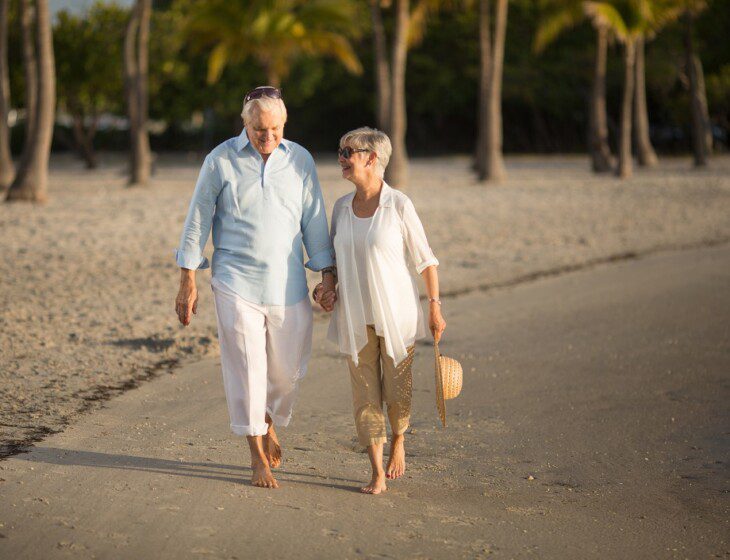 smiling senior couple holds hands and walks facing the camera as they leave the beach