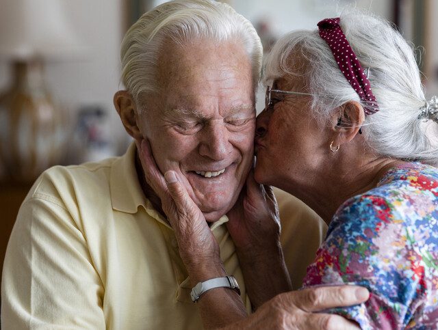 senior woman leans in to kiss the cheek of her smiling husband