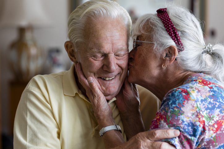 senior woman leans in to kiss the cheek of her smiling husband