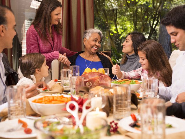 senior woman seated at the head a table smiles and shares a Thanksgiving meal with family