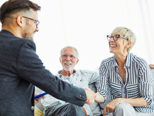 senior woman seated next to her husband on a couch smiles while shaking the hand of a financial counselor