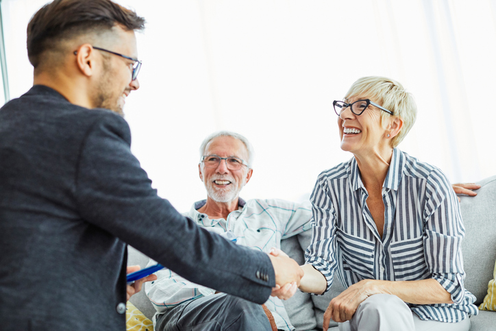 senior woman seated next to her husband on a couch smiles while shaking the hand of a financial counselor