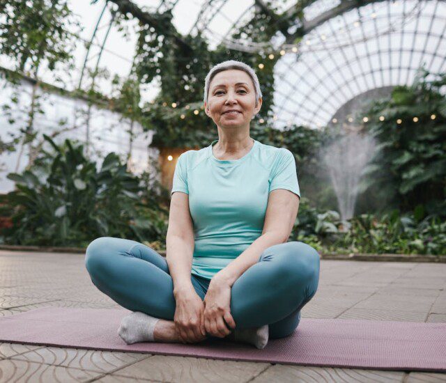 senior woman sits on a yoga mat inside of a greenhouse, smiling
