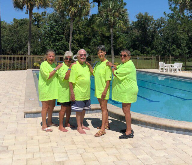 senior residents in matching green shirts stand in front of a resort-style swimming pool at Village on the Green