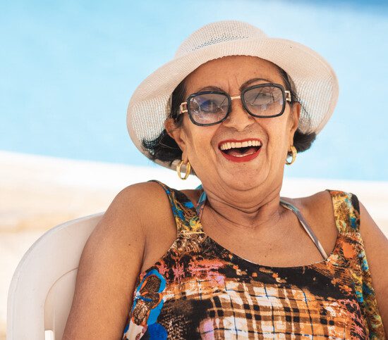 laughing senior woman in beach attire and hat sits in chair at the beach