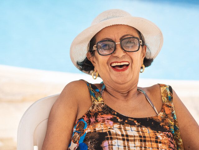 laughing senior woman in beach attire and hat sits in chair at the beach