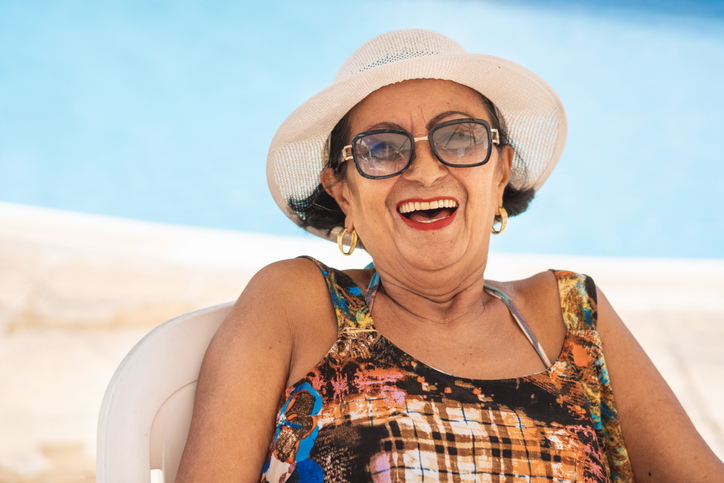 laughing senior woman in beach attire and hat sits in chair at the beach