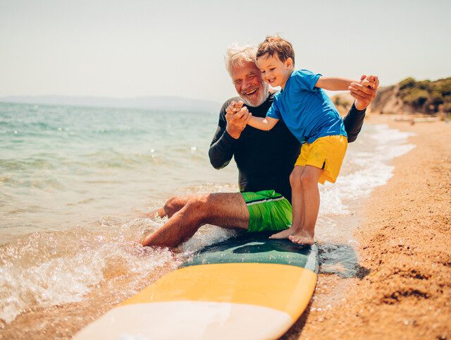 senior man teaches his toddler grandson how to stand on a surfboard on the sand at the beach