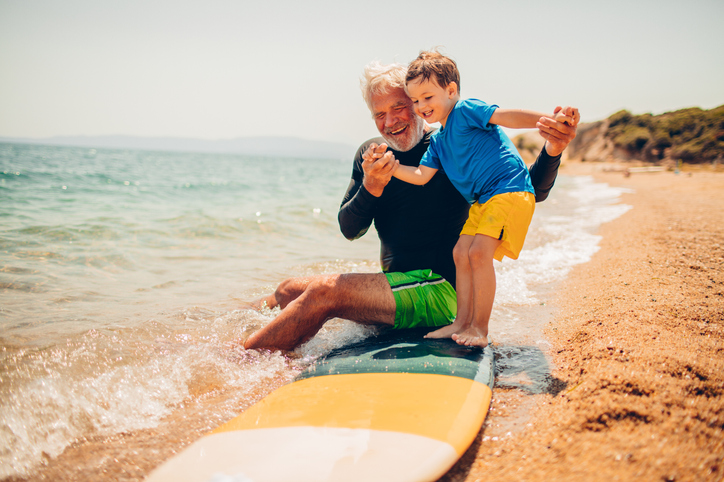 senior man teaches his toddler grandson how to stand on a surfboard on the sand at the beach