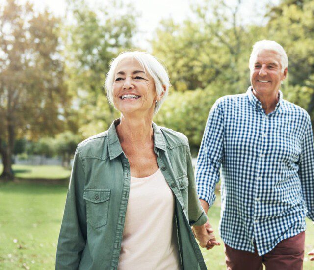 senior woman walks forward, sun on her face, with her senior husband holding her hand and following close behind, both smiling