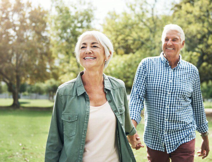 senior woman walks forward, sun on her face, with her senior husband holding her hand and following close behind, both smiling