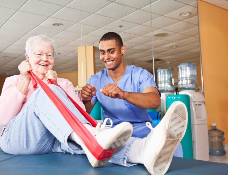 senior woman uses resistance band to complete physical therapy exercise as her trainer excitedly encourages her