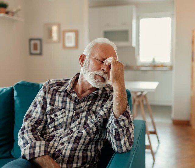 senior man sitting on teal couch pinches the bridge of his nose with his finger and thumb, distressed