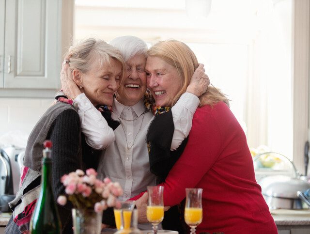 group of three senior women with mimosas smile and embrace