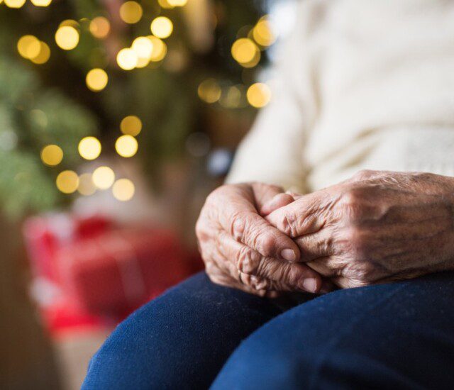 close-up of senior woman's hands folded on her lap, a blurred and illuminated Christmas tree behind her