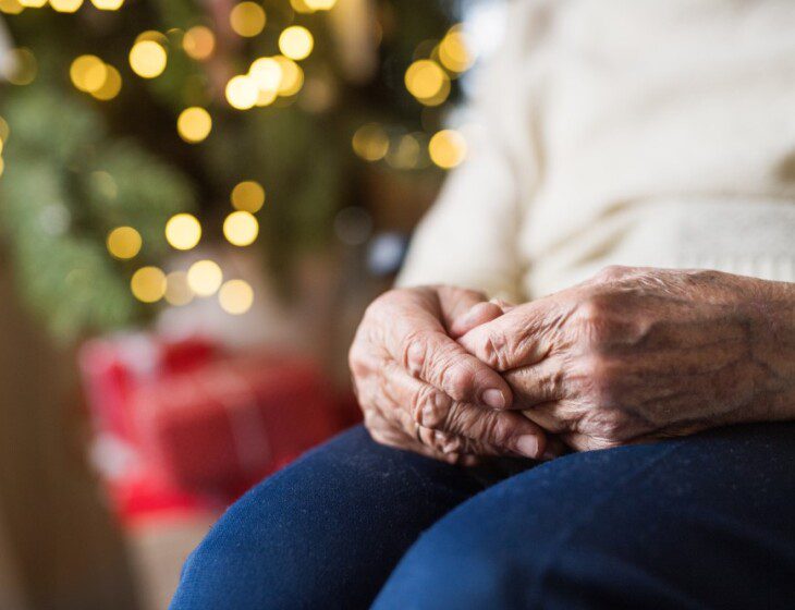 close-up of senior woman's hands folded on her lap, a blurred and illuminated Christmas tree behind her