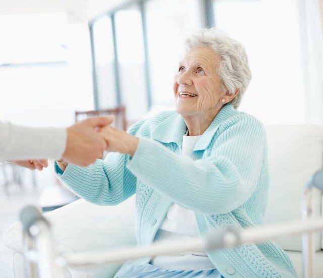 seated senior woman with walker smiles up at her female caregiver, who reaches down to help her stand