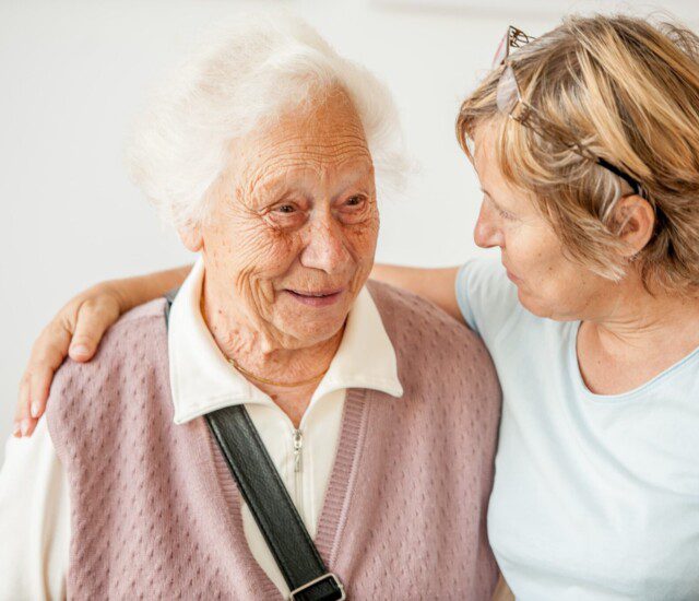 seated senior woman smiles as her adult daughter wraps an arm around her shoulders