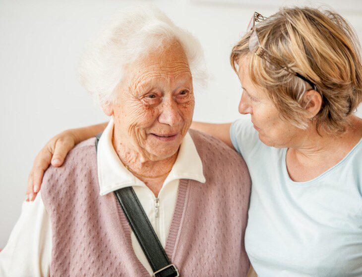 seated senior woman smiles as her adult daughter wraps an arm around her shoulders