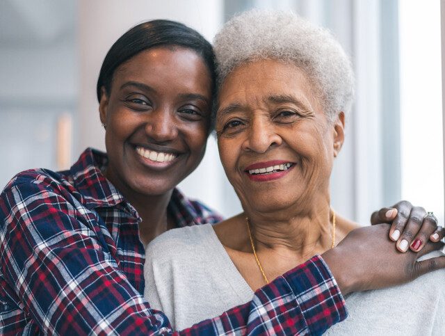 senior woman stands with her adult daughter, whose arms are wrapped around her shoulders, both smiling